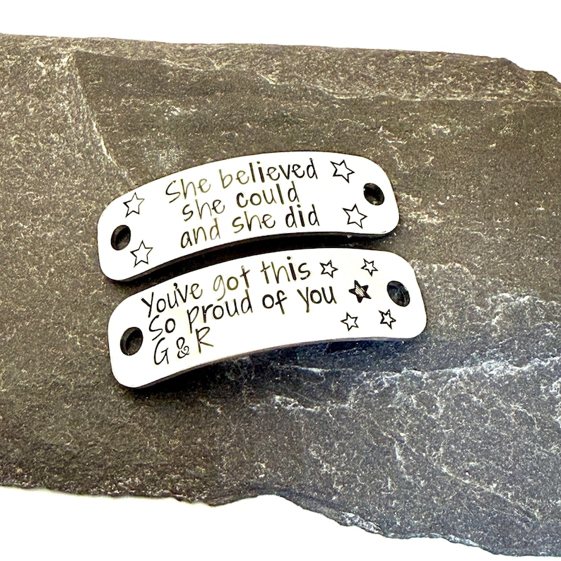 May include: Two silver-colored metal tags with inspirational quotes and star designs. The top tag reads "She believed she could and she did." The bottom tag says "You've got this. So proud of you G&R."