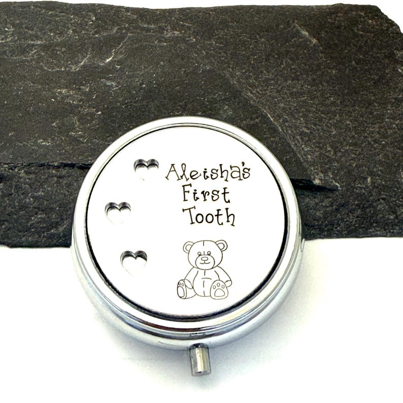 Puede incluir: Una caja redonda plateada con la inscripci&oacute;n "Aleisha's First Tooth" y una ilustraci&oacute;n de un osito de peluche. Tres recortes en forma de coraz&oacute;n est&aacute;n en el lado izquierdo de la tapa. La caja est&aacute; sobre una superficie gris oscuro.