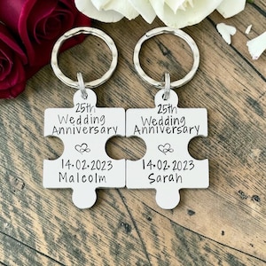 May include: Two silver-toned metal puzzle piece keychains with circular rings. Each piece is engraved with the text 25th Wedding Anniversary, a heart infinity symbol, the date 14.02.2023, and the names Malcolm on one and Sarah on the other.