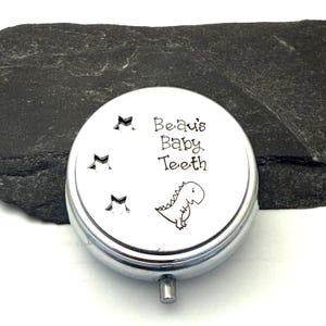 Puede incluir: Una peque&ntilde;a caja redonda plateada para guardar dientes de beb&eacute;. La tapa presenta el texto "Beau's Baby Teeth" con una ilustraci&oacute;n de dinosaurio de dibujos animados y tres recortes de estrellas. La caja tiene un peque&ntilde;o cierre rectangular.