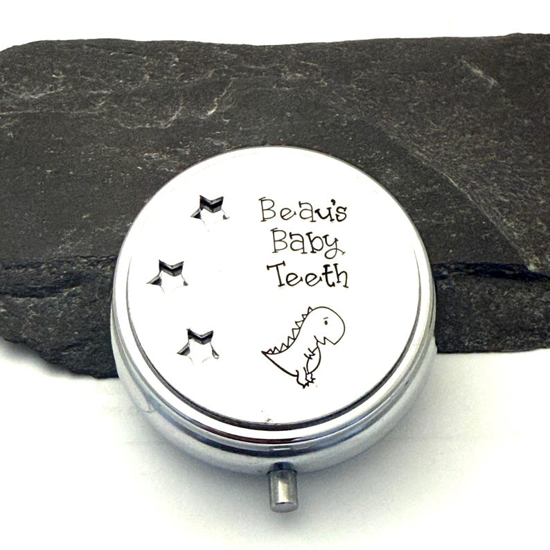Puede incluir: Una peque&ntilde;a caja redonda plateada para guardar dientes de beb&eacute;. La tapa presenta el texto "Beau's Baby Teeth" con una ilustraci&oacute;n de dinosaurio de dibujos animados y tres recortes de estrellas. La caja tiene un peque&ntilde;o cierre rectangular.