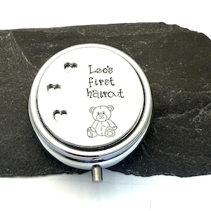 May include: A silver-colored, round keepsake box with the text "Leo's first haircut" and a teddy bear illustration. Three heart-shaped cutouts are on the left side of the lid. The box has a small, cylindrical handle.