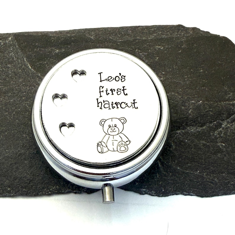 May include: A silver-colored, round keepsake box with the text "Leo's first haircut" and a teddy bear illustration. Three heart-shaped cutouts are on the left side of the lid. The box has a small, cylindrical handle.