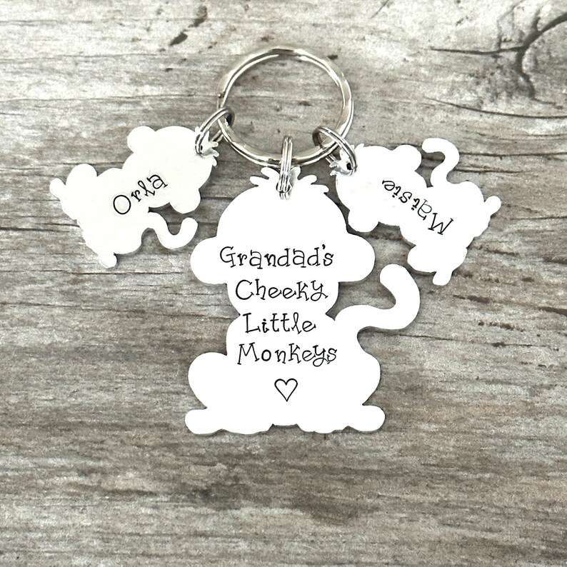 Puede incluir: Llavero de mono plateado con tres dijes. El dije central dice "Granddad's Cheeky Little Monkeys" con un s&iacute;mbolo de coraz&oacute;n. Los otros dos dijes dicen "Orla" y "Mews".