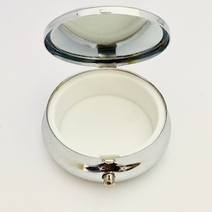 May include: Open, silver-colored pill box with a hinged lid and a white interior. The pill box is round and has a small clasp to secure the lid. The lid has a mirror on the inside.