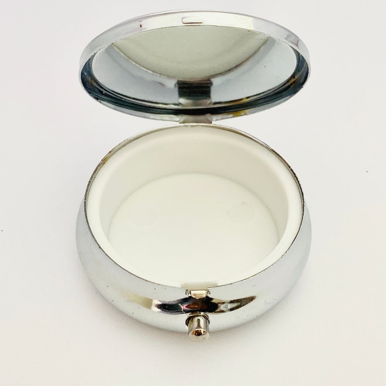 May include: Open, silver-colored pill box with a hinged lid and a white interior. The pill box is round and has a small clasp to secure the lid. The lid has a mirror on the inside.