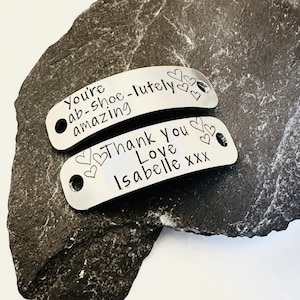 May include: Two curved, silver-toned metal tags with holes at each end. The top tag is engraved with the text "You're ab-shoe-lutely amazing" and three small hearts. The bottom tag is engraved with "Thank you Love Isabelle xxx" and four hearts.