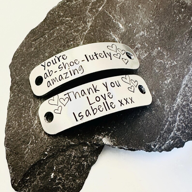 May include: Two curved, silver-toned metal tags with holes at each end. The top tag is engraved with the text "You're ab-shoe-lutely amazing" and three small hearts. The bottom tag is engraved with "Thank you Love Isabelle xxx" and four hearts.