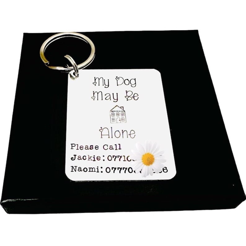 May include: White metal dog tag keyring with the text "My Dog May Be Alone" and a house icon. The tag also includes the text "Please Call" and two phone numbers: Jackie: 07710 and Naomi: 077706. A white daisy with a yellow centre is on the right side of the tag.