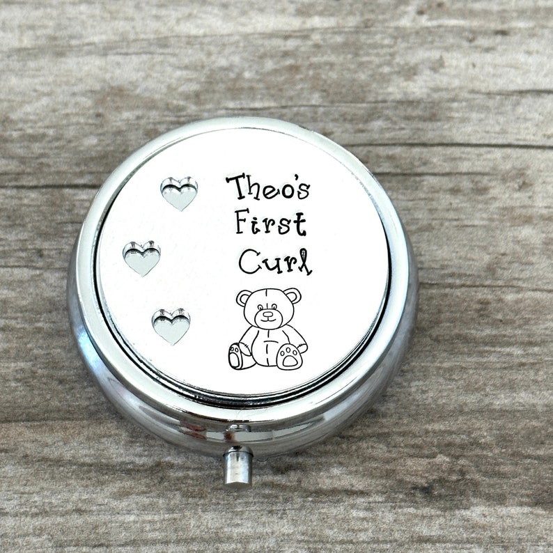 May include: A round, silver-colored keepsake box with the text "Theo's First Curl" and a small teddy bear illustration. Three heart-shaped cutouts are visible on the top. The box is designed to hold a baby's first curl.