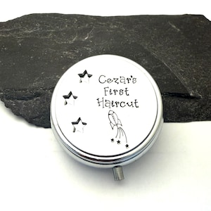 May include: A silver-colored, round keepsake box with the text "Cezar's First Haircut" and a rocket ship illustration. The box has three star cutouts and a small, silver knob. The background is a dark, textured surface.