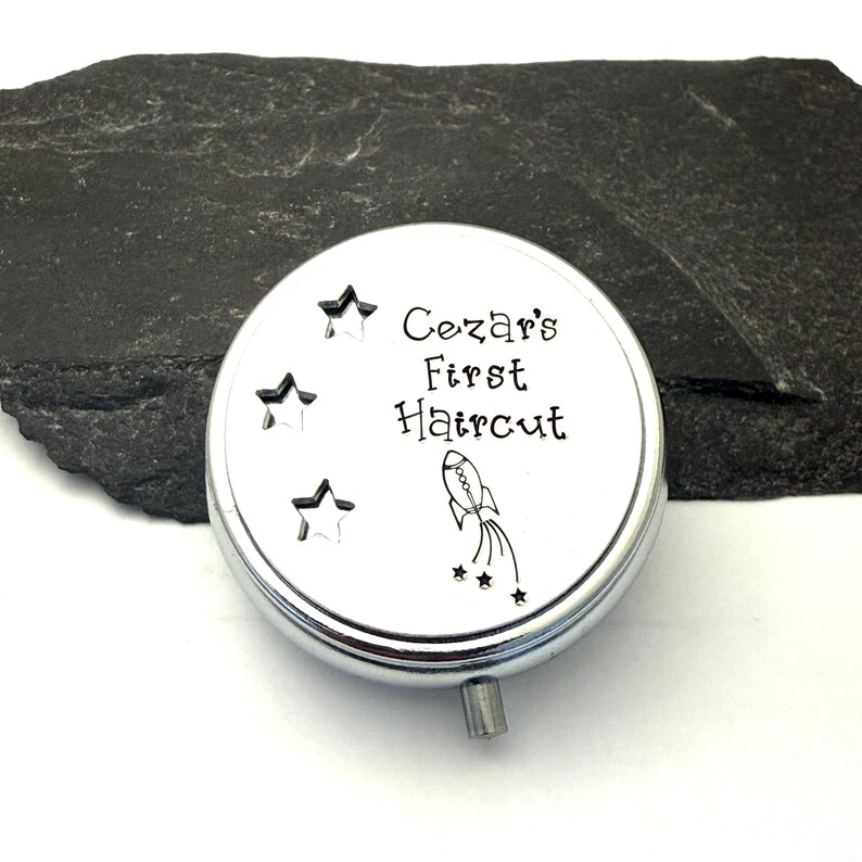 May include: A silver-colored, round keepsake box with the text "Cezar's First Haircut" and a rocket ship illustration. The box has three star cutouts and a small, silver knob. The background is a dark, textured surface.
