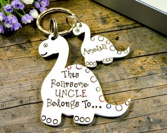 May include: A silver-toned metal keychain featuring two dinosaur-shaped charms attached to a circular ring. The larger dinosaur is engraved with the text "This Roarsome UNCLE Belongs To..." and the smaller dinosaur is engraved with the name "Amelia".