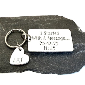 Puede incluir: Un llavero plateado con una etiqueta rectangular y un dije en forma de coraz&oacute;n. La etiqueta rectangular tiene el texto "It Started With A Message..." y la fecha y hora "23.12.25 11:45". El dije de coraz&oacute;n tiene las iniciales "A & C".