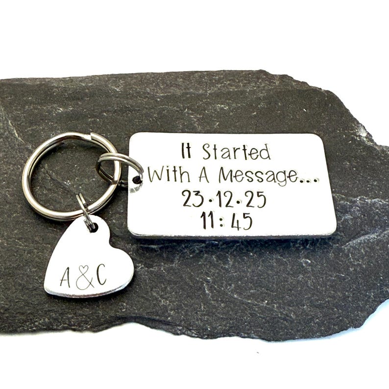 Puede incluir: Un llavero plateado con una etiqueta rectangular y un dije en forma de coraz&oacute;n. La etiqueta rectangular tiene el texto "It Started With A Message..." y la fecha y hora "23.12.25 11:45". El dije de coraz&oacute;n tiene las iniciales "A & C".