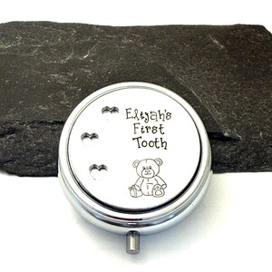 Puede incluir: Una caja redonda de recuerdo de color plateado con el texto "Elijah's First Tooth" y una peque&ntilde;a ilustraci&oacute;n de un oso de peluche. La caja tiene tres recortes en forma de coraz&oacute;n y un peque&ntilde;o asa plateada.