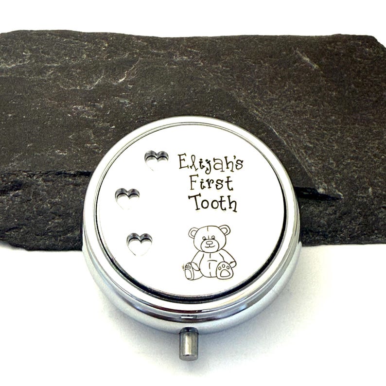 Puede incluir: Una caja redonda de recuerdo de color plateado con el texto "Elijah's First Tooth" y una peque&ntilde;a ilustraci&oacute;n de un oso de peluche. La caja tiene tres recortes en forma de coraz&oacute;n y un peque&ntilde;o asa plateada.