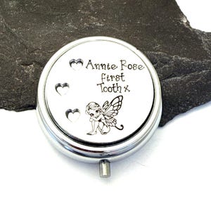 Puede incluir: Una caja redonda de recuerdo de color plateado con tapa abisagrada. La tapa presenta el texto "Annie Rose first Tooth x" y tres recortes en forma de coraz&oacute;n. Tambi&eacute;n hay una peque&ntilde;a ilustraci&oacute;n de un hada.