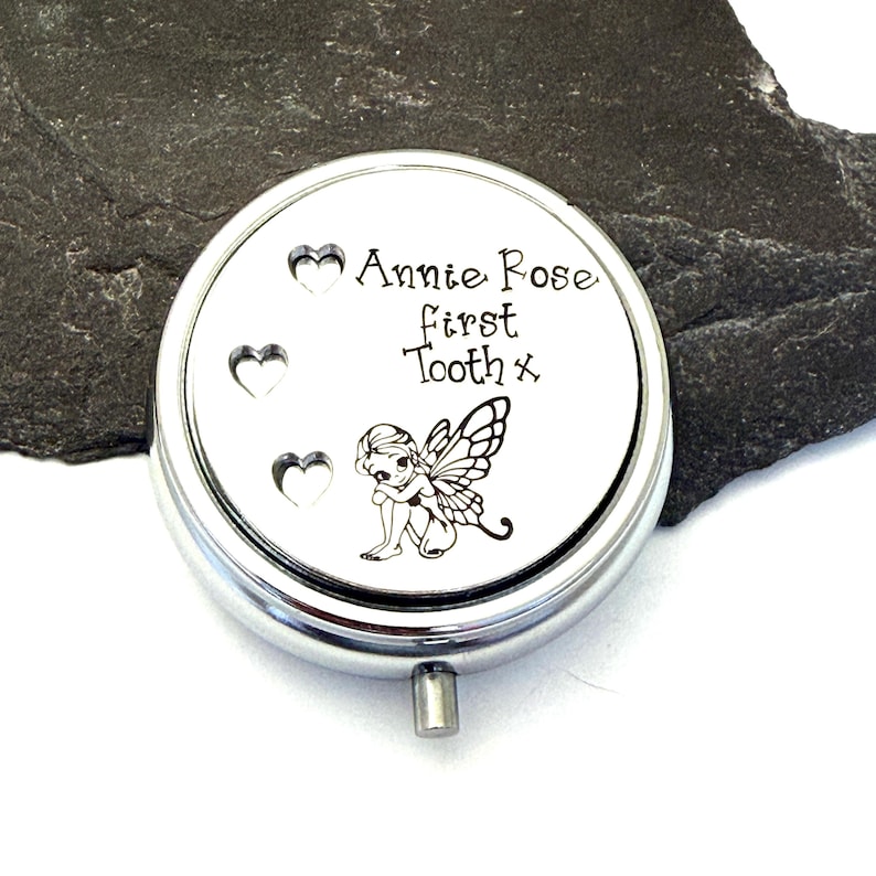 Puede incluir: Una caja redonda de recuerdo de color plateado con tapa abisagrada. La tapa presenta el texto "Annie Rose first Tooth x" y tres recortes en forma de coraz&oacute;n. Tambi&eacute;n hay una peque&ntilde;a ilustraci&oacute;n de un hada.