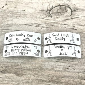 May include: Four rectangular silver-toned metal tags with rounded edges and two holes for attachment. The tags feature engraved black text and graphics including running shoes, stars, and figures of people running, with personalized names.