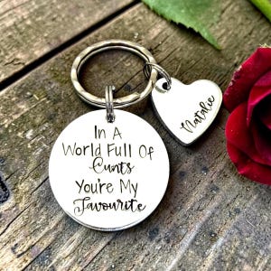 May include: A silver-toned metal keyring featuring a circular disc engraved with the text In A World Full Of Cunts You're My Favourite and a smaller heart-shaped charm engraved with the name Natalie, both attached to a split ring.
