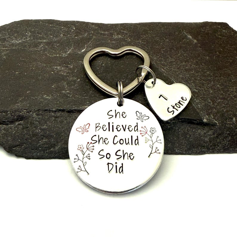 Pode incluir: Um porta-chaves de metal prateado com um anel em forma de cora&ccedil;&atilde;o e um pendente redondo gravado com o texto She Believed She Could So She Did, acompanhado de motivos florais e borboletas e um pequeno cora&ccedil;&atilde;o com 1 Stone.
