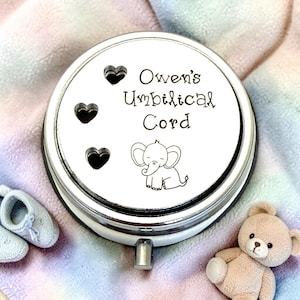 May include: A silver-colored, round keepsake box with the text "Owen's Umbilical Cord" and a small elephant illustration. The box has three heart-shaped cutouts and a small winding key. A teddy bear and baby shoes are in the background.