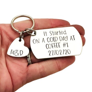 Puede incluir: Un llavero plateado con un dije en forma de coraz&oacute;n grabado con "M&D" y una etiqueta rectangular. La etiqueta dice "It Started ON A COLD DAY AT COFFEE #1 27/02/20" en texto negro. El llavero se sostiene en una mano.