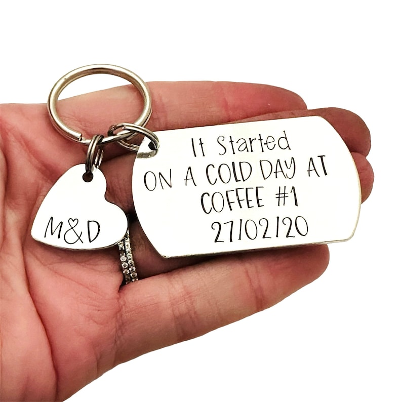 Puede incluir: Un llavero plateado con un dije en forma de coraz&oacute;n grabado con "M&D" y una etiqueta rectangular. La etiqueta dice "It Started ON A COLD DAY AT COFFEE #1 27/02/20" en texto negro. El llavero se sostiene en una mano.