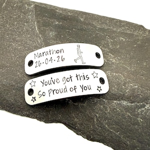 May include: Two rectangular silver-toned metal shoe tags with rounded edges and holes on each side. The top tag features the text Marathon 26.04.26 and a runner icon. The bottom tag features the text You've got this So Proud of You with stars.