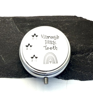 Puede incluir: Una caja plateada para guardar dientes de beb&eacute; con el texto "Harvey's Baby Teeth" en la tapa, junto con ilustraciones de estrellas y arco&iacute;ris. La caja se muestra sobre una superficie oscura y texturizada.