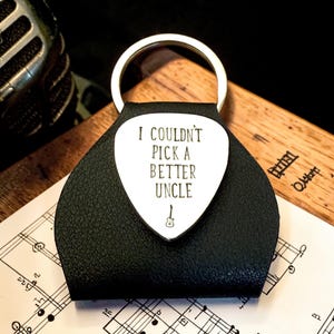 May include: A black leather guitar pick holder with a silver metal guitar pick attached to a circular keyring. The pick is engraved with the text I COULDN'T PICK A BETTER UNCLE and a small guitar icon centred at the bottom of the inscription.