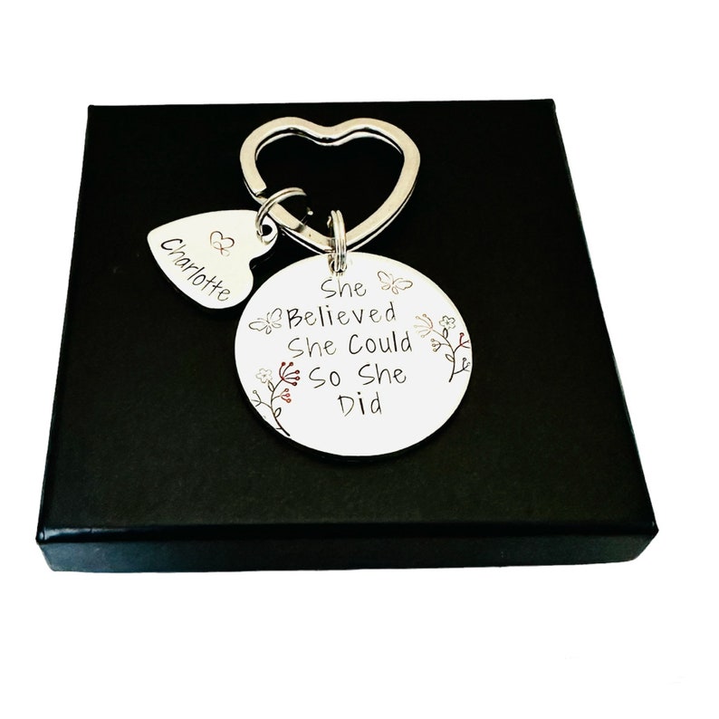 Pode incluir: Um porta-chaves de metal prateado com um anel em forma de cora&ccedil;&atilde;o, um pequeno pendente em forma de cora&ccedil;&atilde;o gravado com o nome Charlotte e um disco circular maior gravado com o texto She Believed She Could So She Did, decorado com motivos florais e borboletas.