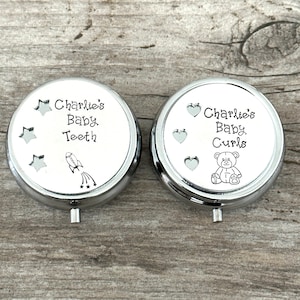 May include: Two silver-colored, round keepsake boxes with white tops. One box reads "Charlie's Baby Teeth" with star and rocket ship illustrations. The other reads "Charlie's Baby Curls" with heart and teddy bear illustrations.