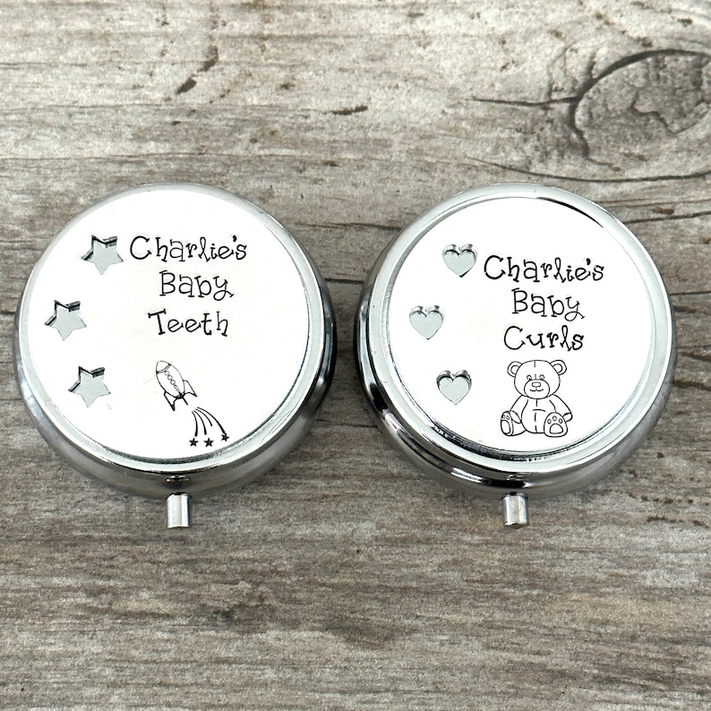 May include: Two silver-colored, round keepsake boxes with white tops. One box reads "Charlie's Baby Teeth" with star and rocket ship illustrations. The other reads "Charlie's Baby Curls" with heart and teddy bear illustrations.