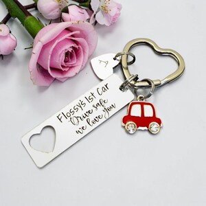 May include: A silver keychain with a heart-shaped ring and a rectangular tag with the text "Flossy's 1st Car Drive safe we love you." A red car charm with rhinestone wheels hangs below. A small letter "A" tag is also attached.
