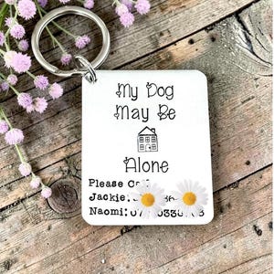 May include: A white rectangular metal keyring with a silver split ring. The text reads: My Dog May Be Alone, followed by a house icon, the word Alone, and contact lines for Jackie and Naomi. The design features small heart accents.