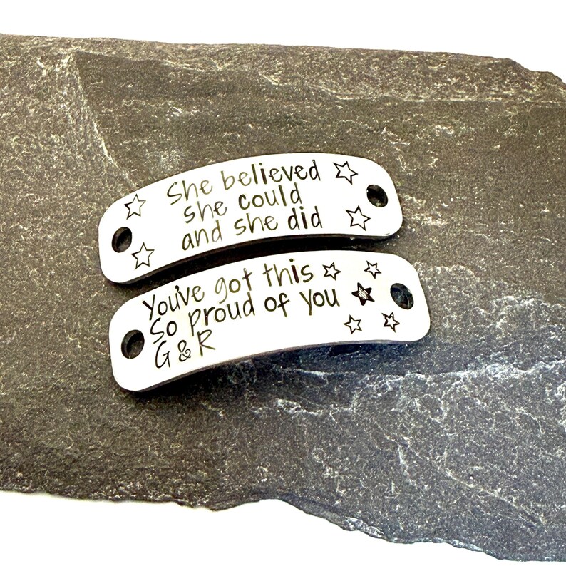 May include: Two curved silver-toned metal tags with holes on each end. The top tag is engraved with the text "She believed she could and she did" and star icons. The bottom tag features the text "You've got this So proud of you G&R" and stars.