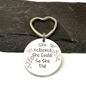 Pode incluir: Um porta-chaves redondo de metal prateado com um anel em forma de cora&ccedil;&atilde;o. A superf&iacute;cie est&aacute; gravada com o texto "She Believed She Could So She Did" e decorada com pequenas ilustra&ccedil;&otilde;es de borboletas e ramos florais nos lados.