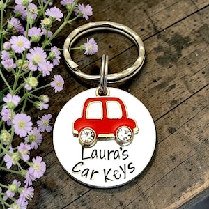 May include: A silver car key ring with a red car charm and the text "Laura's Car Keys". The car charm has clear gem wheels. The key ring is round and has a hammered texture. The background includes purple flowers and a wooden surface.