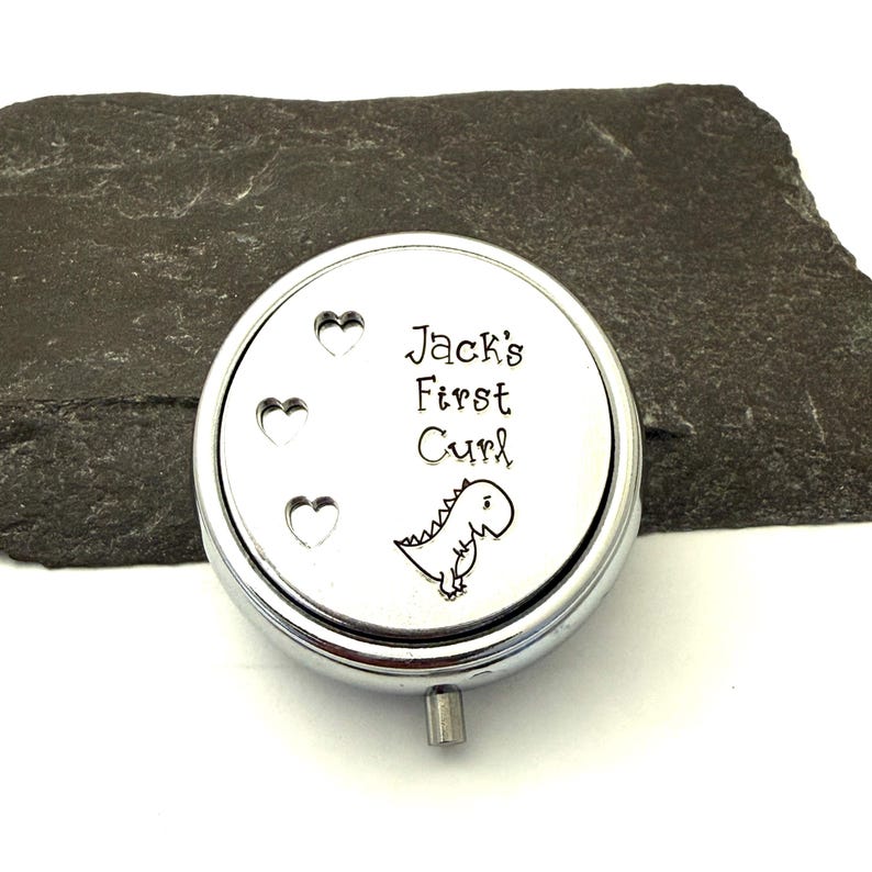 May include: A round, silver-colored keepsake box with the text "Jack's First Curl" and a small dinosaur illustration. Three heart shapes are etched on the left side of the lid. The box has a small, cylindrical handle.