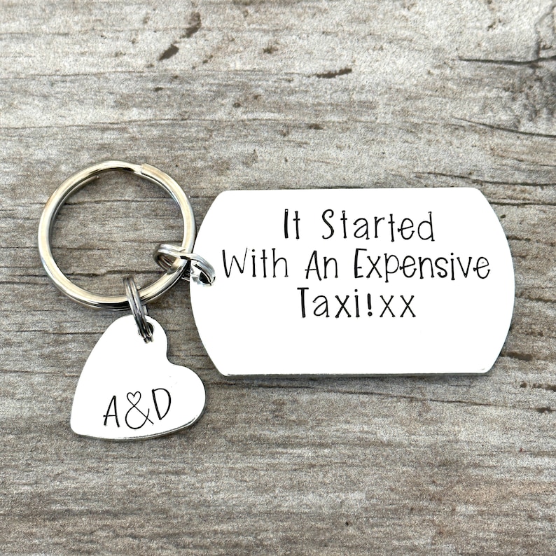 Puede incluir: Un llavero plateado con un dije en forma de coraz&oacute;n y una etiqueta rectangular. El dije de coraz&oacute;n tiene las iniciales "A&D". La etiqueta dice "It Started With An Expensive Taxi!xx".