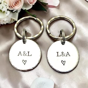 May include: Two silver keyrings with round, hammered-metal pendants. Each pendant has initials and a heart symbol. One keyring reads "A&L" and the other reads "L&A". The keyrings are on a satin surface with flowers in the background.