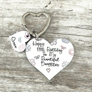 May include: White heart-shaped keychain with a floral pattern and the text "Happy 18th Birthday To My Beautiful Daughter" with a small heart at the bottom. A smaller heart-shaped charm with the name "Grace" is attached to the keychain.