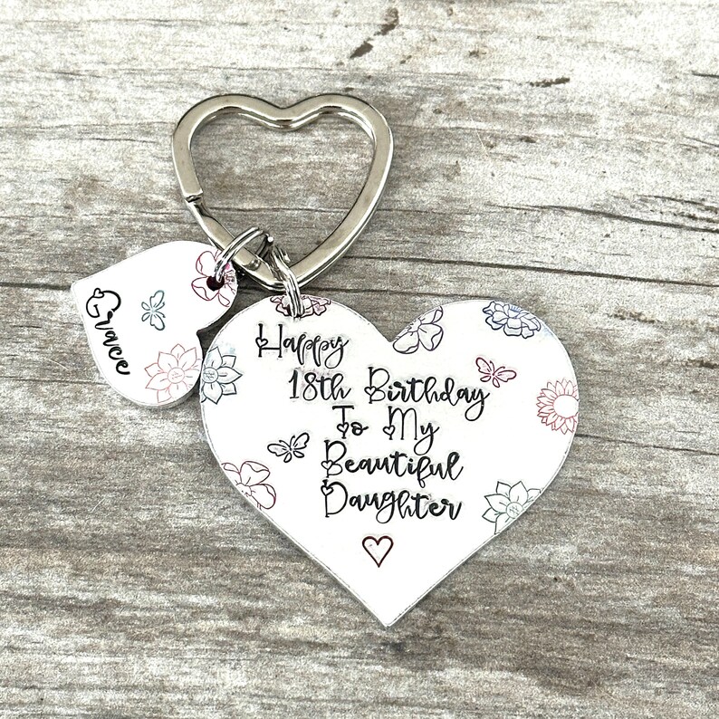 May include: White heart-shaped keychain with a floral pattern and the text "Happy 18th Birthday To My Beautiful Daughter" with a small heart at the bottom. A smaller heart-shaped charm with the name "Grace" is attached to the keychain.