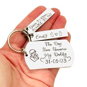 May include: Silver keychain with three tags. The largest tag has a father and child illustration and the text "The Day You Became My Daddy 31.01.23". The middle tag has the name "Emily" and a footprint symbol. The smallest tag has the text "Born 9:48 am, Weight 7lb 5oz".