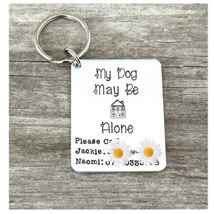 May include: Silver keyring with a white tag that reads "My Dog May Be Alone. Please Call Jackie: 36. Naomi: 070330.3". The tag has a small house icon and two white daisies with yellow centres.