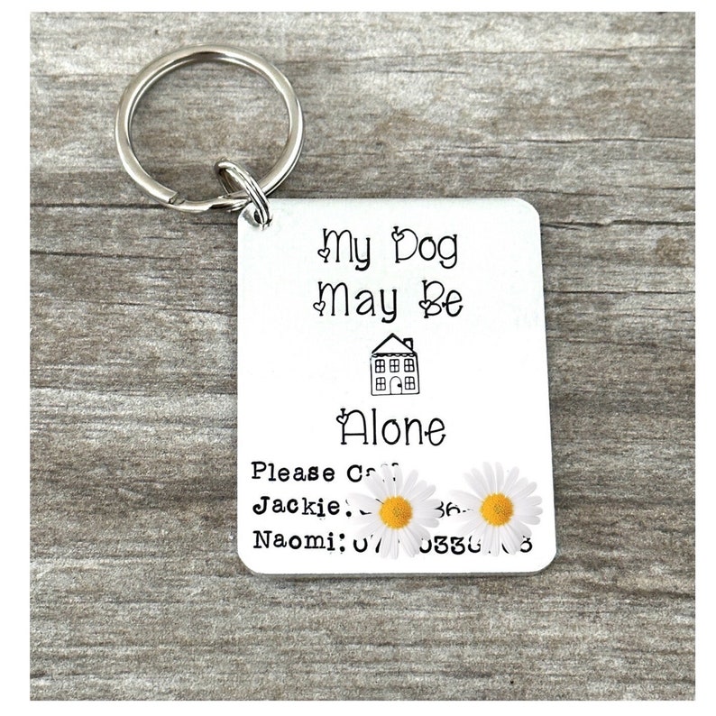 May include: Silver keyring with a white tag that reads "My Dog May Be Alone. Please Call Jackie: 36. Naomi: 070330.3". The tag has a small house icon and two white daisies with yellow centres.
