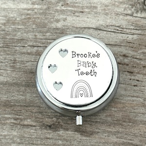 Puede incluir: Una caja redonda plateada para guardar dientes de beb&eacute;. La tapa tiene el texto "Brooke's Baby Teeth" con tres corazones y un dise&ntilde;o de arco&iacute;ris. La caja est&aacute; sobre una superficie de madera.