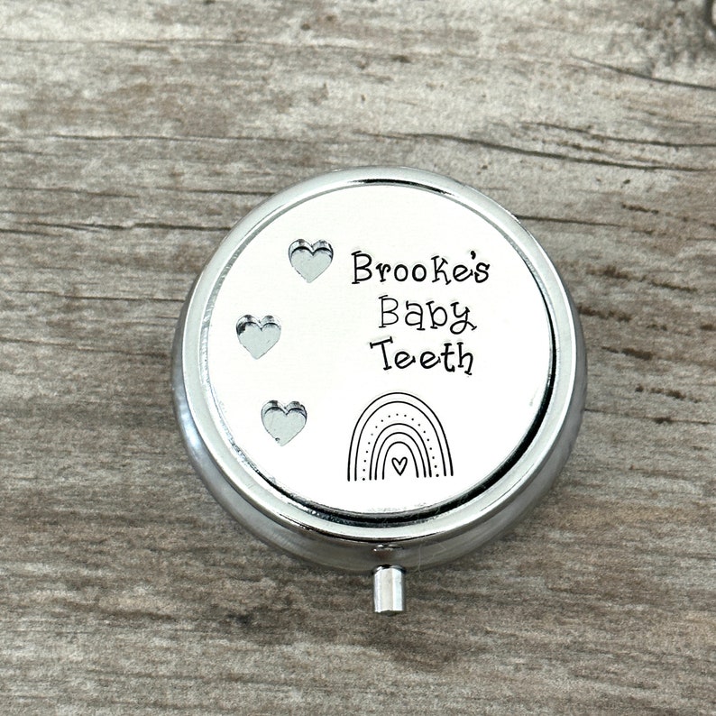 Puede incluir: Una caja redonda plateada para guardar dientes de beb&eacute;. La tapa tiene el texto "Brooke's Baby Teeth" con tres corazones y un dise&ntilde;o de arco&iacute;ris. La caja est&aacute; sobre una superficie de madera.